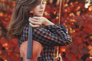 Kırmızı kemanı olan genç bir kadın ve sonbahar doğasının arka planında selam veren romantik bir kız müzikal sanatla meşgul, hobi anlayışı, açık havada konser.