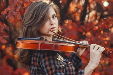 Kırmızı kemanı olan genç bir kadın ve sonbahar doğasının arka planında selam veren romantik bir kız müzikal sanatla meşgul, hobi anlayışı, açık havada konser.