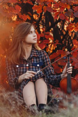 Yerde keman çalan genç bir kadın, arka planda kırmızı sonbahar yaprakları olan bir kemanla, doğada müzik aleti olan romantik bir kız, bir hobi ve sanat konsepti.