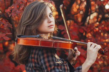 Kırmızı kemanı olan genç bir kadın ve sonbahar doğasının arka planında selam veren romantik bir kız müzikal sanatla meşgul, hobi anlayışı, açık havada konser.