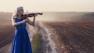 Uzun elbiseli bir kadın tozdan bir kulüp ile arka planda keman çalıyor, müzikal sanatla meşgul, doğada performans, müzikte konsept tutku
