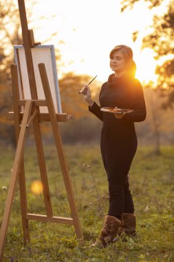 Genç kadın ressamın silueti sehpaya resim çiziyor, arka planda bir fırça ve renk paleti olan bir kız figürü, hobi ve sanat kavramı.
