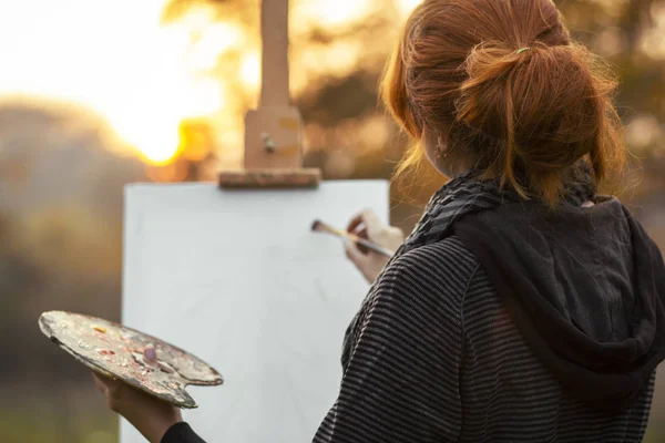 Cabeza trasera de chica pelirroja con un moño pintando un cuadro en un caballete en la naturaleza, una mujer joven dedicada al arte y disfrutando de un hermoso paisaje al atardecer, concepto hobby y estilo de vida — Foto de Stock