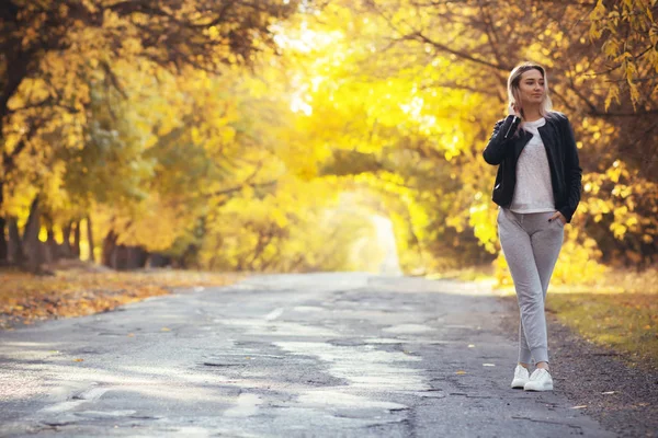 Jovem mulher em sportswear casual com uma mochila em pé na estrada de asfalto de uma estrada suburbana, menina viaja andando e apreciando a natureza do outono — Fotografia de Stock