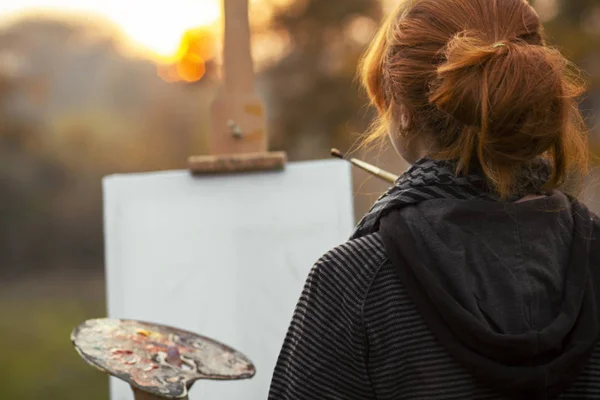 Kızıl saçlı bir kızın arka kafası, bir somun topuzu ile sehpaya resim çiziyor, sanatla ilgilenen ve günbatımında güzel manzaranın tadını çıkaran genç bir kadın, konsept hobisi ve yaşam tarzı — Stok fotoğraf