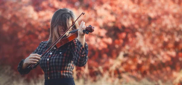 Hermosa joven tocando el violín sobre un fondo de follaje rojo, chica romántica en vestido tocando un instrumento musical en la naturaleza, actuación musical al aire libre, concepto de hobby y pasión en el arte — Foto de Stock