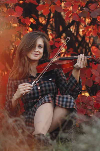 Herfstportret van een mooie vrouw op de grond met een viool onder de kin op een achtergrond van rood gebladerte, meisje bezig met het bespelen van een muziekinstrument in de natuur, een concept van passie in de kunst — Stockfoto