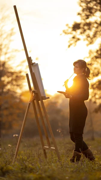 silhouette of young woman artist painting a picture on the easel, girl figure with a brush and a palette of colors on the background of autumn landscape, a concept of hobby and art