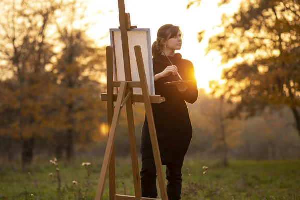 silhouette of young woman artist painting a picture on the easel, girl figure with a brush and a palette of colors on the background of autumn landscape, a concept of hobby and art