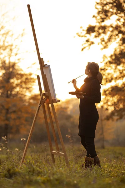 Silueta de artista joven pintando un cuadro en el caballete, figura de niña con un pincel y una paleta de colores en el fondo del paisaje de otoño, un concepto de hobby y arte — Foto de Stock