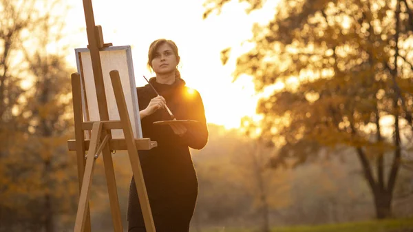 Sziluett fiatal nő művész festmény egy képet a festőállvány, lány alak ecsettel és egy színpaletta a háttérben az őszi táj, a koncepció a hobbi és a művészet — Stock Fotó