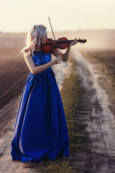 Mujer en vestido largo tocando el violín en el fondo del camino de campo con un club de polvo, chica dedicada al arte musical, actuación en la naturaleza, concepto pasión en la música — Foto de Stock