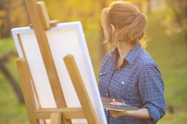 Genç kadın sanatçı bir tablo çiziyor bir sehpaya, fırçalı ve palet boyalı bir kız sonbaharın erken saatlerinden esinlenerek çalışıyor, bir sanat konsepti, bir hobi