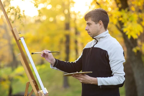 Fiatal művész festett egy képet vászonra egy festőállványra a természetben, egy férfi ecsettel és egy festékpalettával az őszi fák között, egy koncepció a művészeti munka és a hobbi — Stock Fotó