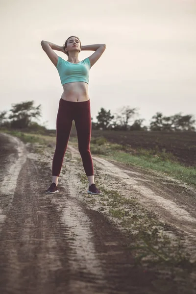 slim young woman in sportswear exercises and stretches hands up in the field, girl engaged in sport outdoors on a cloudy day, concept healthy lifestyle and female beauty