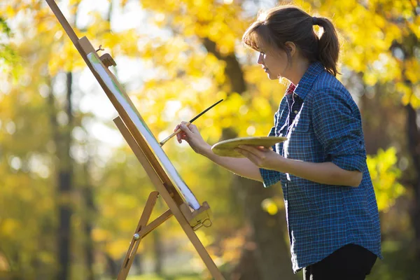 Young woman artist drawing a picture on canvas on an easel in nature, a girl with a brush and a palette of paints working inspired by early autumn, a concept of art, hobby — 스톡 사진