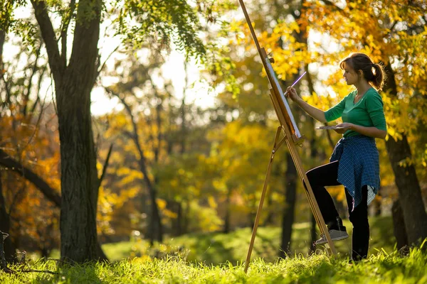 Young woman artist drawing a picture on canvas on an easel in nature, a girl with a brush and a palette of paints working inspired by early autumn, a concept of art, hobby — Stock Photo, Image