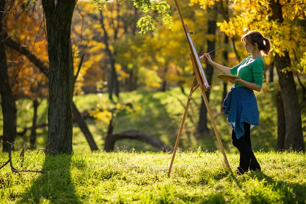 Young woman artist drawing a picture on canvas on an easel in nature, a girl with a brush and a palette of paints working inspired by early autumn, a concept of art, hobby — Stock Photo, Image