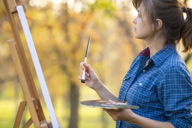 Genç kadın sanatçı bir tablo çiziyor bir sehpaya, fırçalı ve palet boyalı bir kız sonbaharın erken saatlerinden esinlenerek çalışıyor, bir sanat konsepti, bir hobi