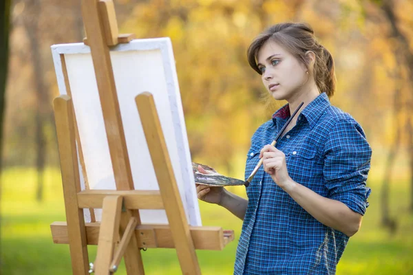 Fiatal nő művész rajz egy képet vászonra egy festőállvány a természetben, egy lány ecsettel és egy paletta festékek munka ihlette kora ősszel, a koncepció a művészet, hobbi — Stock Fotó