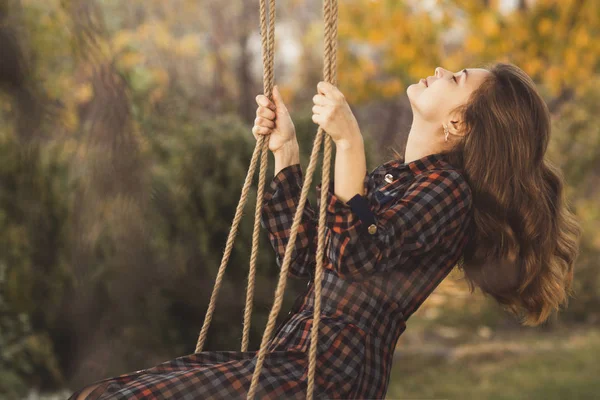 cute girl in a dress sways on a swing in the autumn garden, a young woman in a positive romance mood relaxing on nature