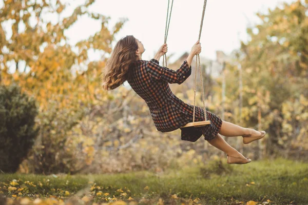 cute girl in a dress sways on a swing in the autumn garden, a young woman in a positive romance mood relaxing on nature