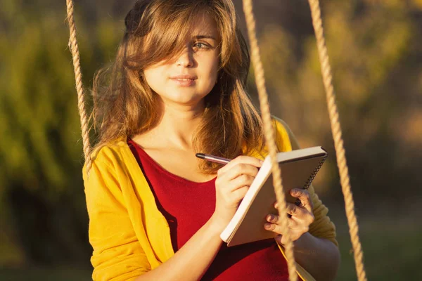 Cute girl on a swing with a notebook and pen keeps a diary of feelings in nature, a woman composes and writes, romantic mood — Stock Photo, Image