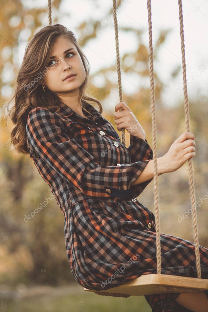 upset girl in a dress sways on a swing in the autumn garden, a young woman in melancholy mood thinkng on nature