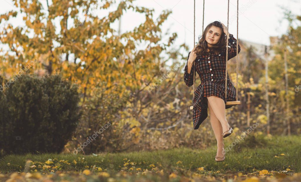 cute girl in a dress sways on a swing in the autumn garden, a young woman in a positive romance mood relaxing on nature