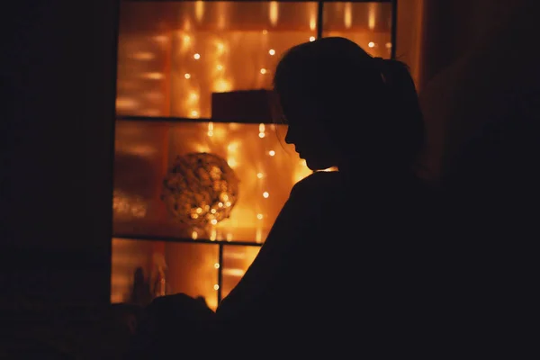 Perfil silhueta figura jovem mulher sentado em uma sala decorada com uma grinalda luz contra prateleiras com caixas de presente e decoração, conceito de relaxamento à noite, humor e solidão — Fotografia de Stock
