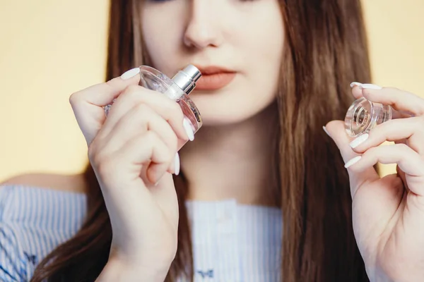 perfume bottle in the hands of a beautiful girl on a studio color background, part of the face of a young woman sniffing aroma, concept of female care and cosmetics