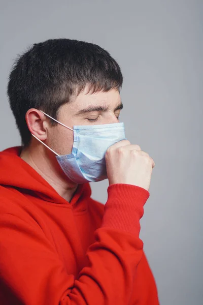 Man in medical mask coughing on gray studio background, concept of prevention viral deseases, person in red sweatshirt with breathing problems