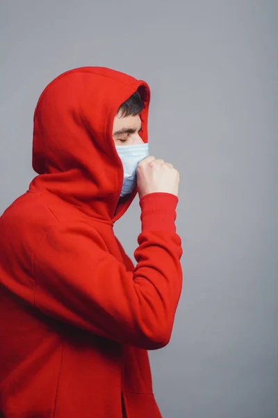 Man in medical mask coughing on gray studio background, concept of prevention viral deseases, person in red sweatshirt with breathing problems