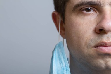 Gri stüdyo geçmişi, konsept tıbbı ve sağlık alanındaki klinikteki zorlu ameliyattan sonra kulaklarında koruyucu maske olan yorgun genç doktorun yüzünün yarısını kapatın.