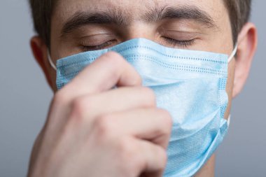 Koruyucu tıbbi maskeli, gri stüdyo geçmişi, konsept tıp ve sağlık üzerine meditasyon yapan yorgun genç doktorun yüzü