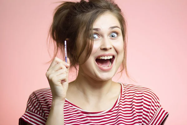 young happy woman with positive result result of a one-time pregnancy test, excited future mother, family planning, motherhood concept on pink studio background