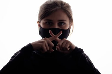 Siyah koruyucu maskeli genç bir kadının silueti. Ağzında iki parmağıyla bir sessizlik jesti gerçeğin sessizliği, bilgi gizliliği, beyaz soyutlanmış arka planda komplo.