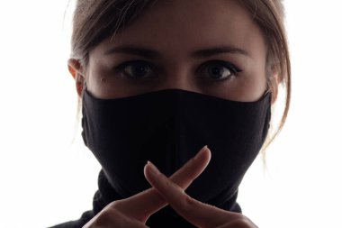 Siyah koruyucu bir maske takan genç bir kadın sessiz bir jest yapıyor. Ağzında iki parmağı çapraz, gerçeğin sessizliği kavramı, gizli bilgiler.