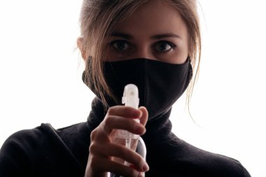 Siyah maskeli genç bir kadın beyaz arka planda dezenfektan spreyi tutuyor, enfeksiyonun yayılması, sağlık güvenlik ajanı, kısırlık ve hijyen kavramına karşı mücadele ediyor.