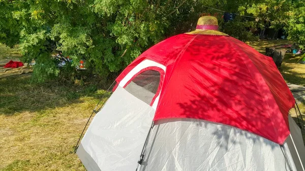 Red and white tent for tourism, camping, recreation for your travel Agency. A small tent in the forest near the lake. Sunny weather. Under the trees, clothes and things of tourists on vacation.