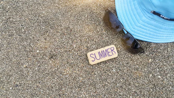 Mavi bir şapka, güneş gözlüğü ve üzerinde yaz yazan bir ağaç. Deniz kumu üzerinde kompozisyon. Yaz havası. Seyahat acenteleri için plaj fotoğrafı. Güneş ışını. Boşluk için metin.