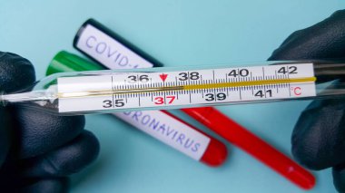 Doktorun elinde yüksek ateş olduğunu gösteren bir termometre. Arka planda, odaklanmadan, kanla dolu iki vakum tüpü var. Coronavirus ve covid-19 metni. Enfeksiyon kavramı.