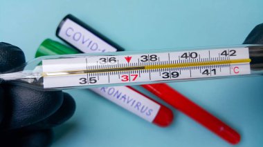 Doktorun elinde yüksek ateş olduğunu gösteren bir termometre. Arka planda, odaklanmadan, kanla dolu iki vakum tüpü var. Coronavirus ve covid-19 metni. Enfeksiyon kavramı.