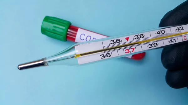 Termometre doktorun elinde yüksek ateş olduğunu gösteriyor. Arka planda odaklanma olmadan içinde kan olan bir vakum tüpü var. Koronavirüs metni. Enfeksiyon kavramı.