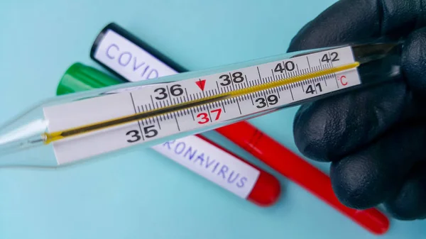 A thermometer showing a high temperature in the doctor\'s hand. In the background, without focus, there are two vacuum tubes with blood. Text of coronavirus and covid-19. The concept of infection.