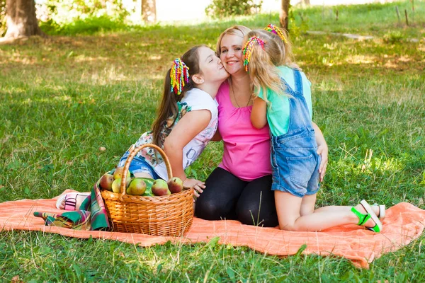 快乐的妈妈带着两个女儿坐在公园里的橙色野餐毯上 女孩亲吻妈妈 — 图库照片