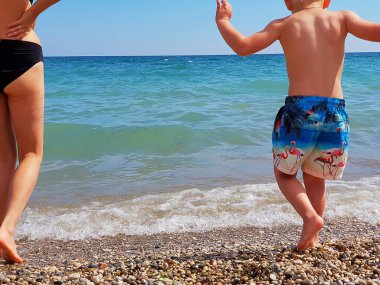 Aile yüzmek istiyor. Anne ve bebek suyun kenarında durur ve denize bakar.