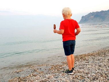 Yazın deniz kıyısında bir çocuk. Çocuk sahilde dondurma yiyor ve sörf sırasına bakıyor..