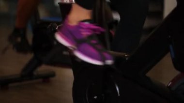 Modern karanlık bir spor salonunda spor aletleri kullanan atletik bir kız. Bisiklet pedalında spor yapmak. Kadın bacakları pedala yakın çekimde. Sağlıklı yaşam tarzı ve spor kavramları. Bisikletli kadın 