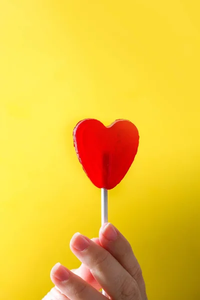Hand roter Lutscher mit Herzform auf gelbem Hintergrund. Liebe. Valentinstag. — Stockfoto